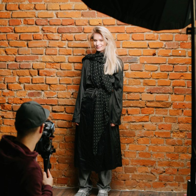 Fashion model in black dress posing against brick wall in studio setting.